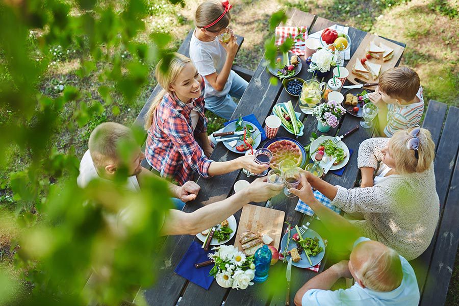 About Our Agency - Family Having Dinner Outdoors at a Picnic Table, Seen From Above Between Tree Branches